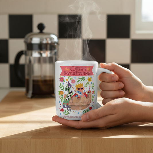 Person holding a white Mary Engelbreit The Queen of Everything 15 oz mug with a whimsical floral design and text, steam rising, in a kitchen setting.