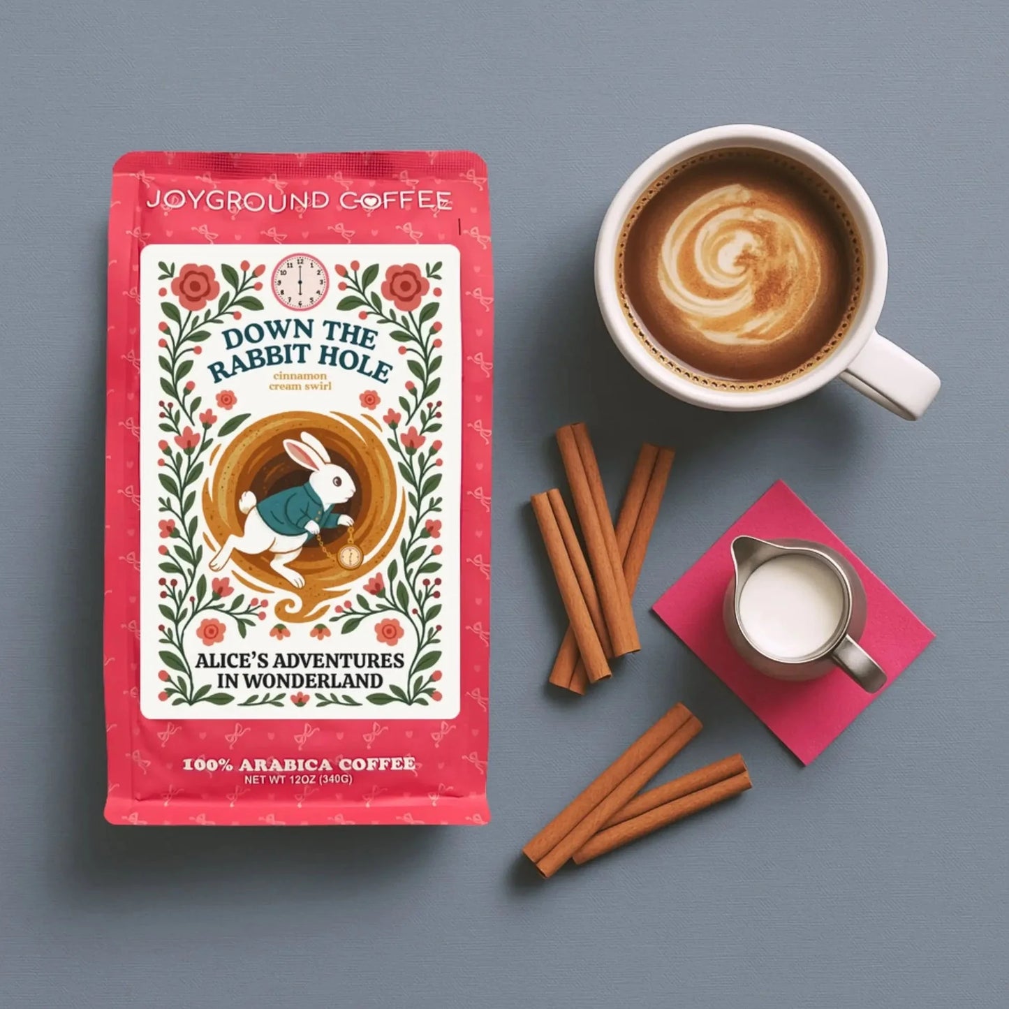 Down The Rabbit Hole pink bag from Joyground Coffee next to a white coffee mug, a pitcher of cream, and cinnamon sticks.