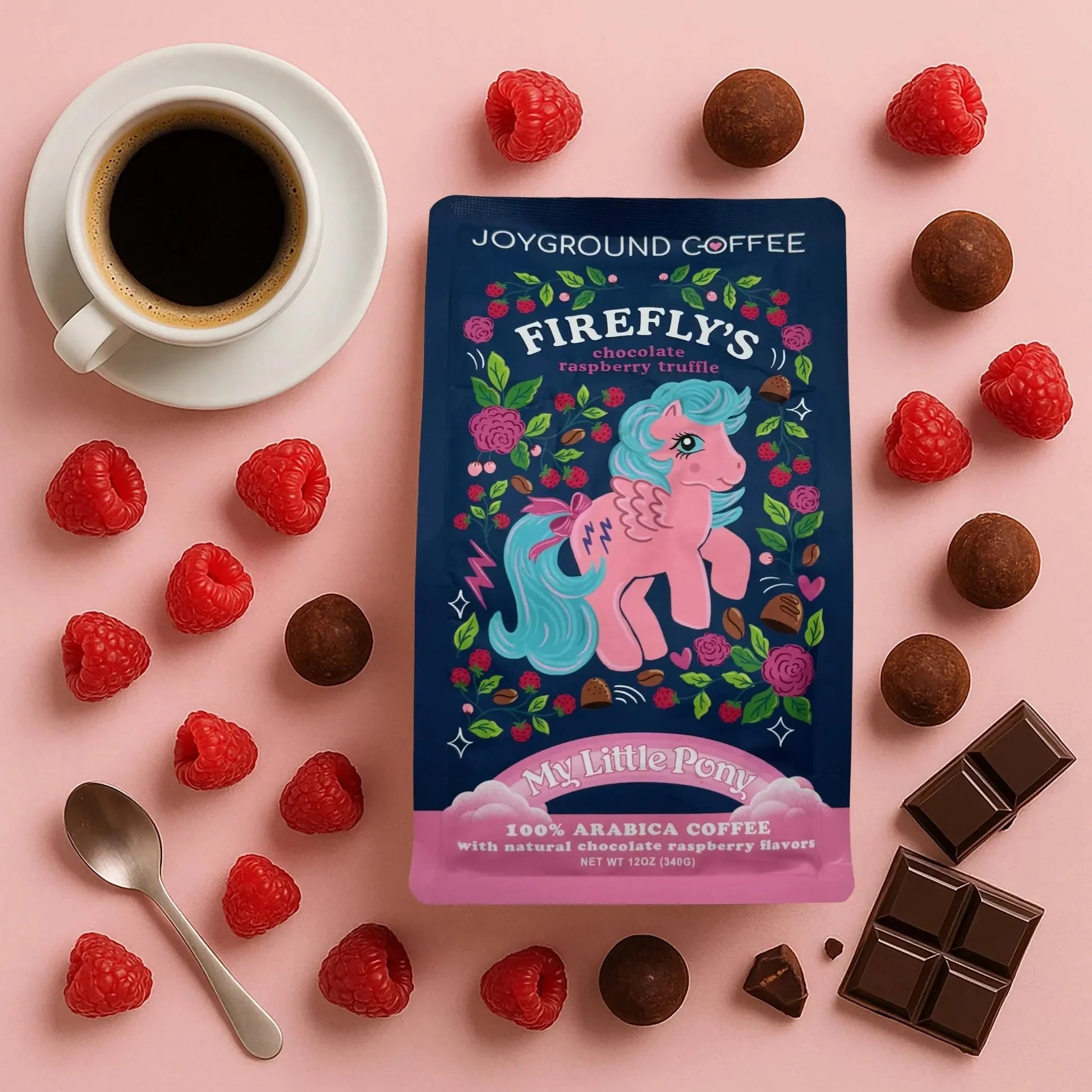 Firefly's Chocolate Raspberry Truffle bag from Joyground Coffee on a pink surface next to rasberries, truffles, a white coffee mug and saucer, and chocolate pieces.