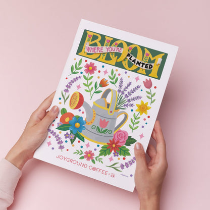 Joyground Coffee x Mary Engelbreit Bloom Where You're Planted kitchen art print held against a pink background.