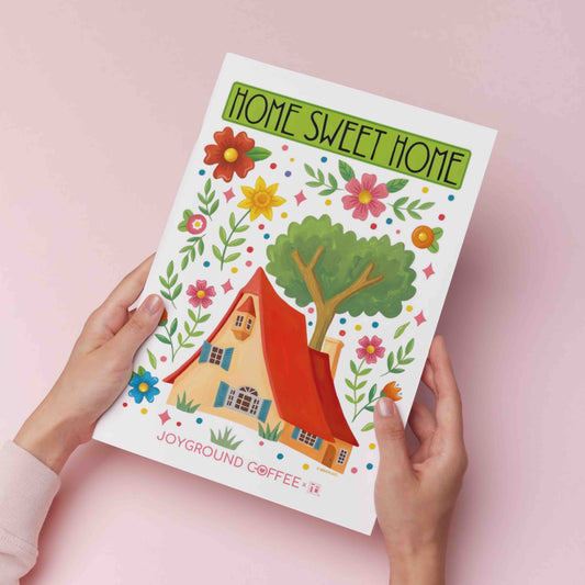 Joyground Coffee x Mary Engelbreit art print titled 'Home Sweet Home' held in hands on a pink background