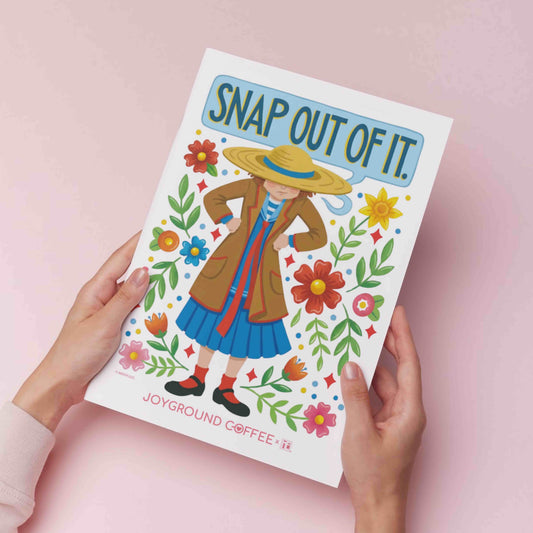 Mary Engelbreit kitchen decor art print titled 'Snap Out of It' held by hands on a pink background